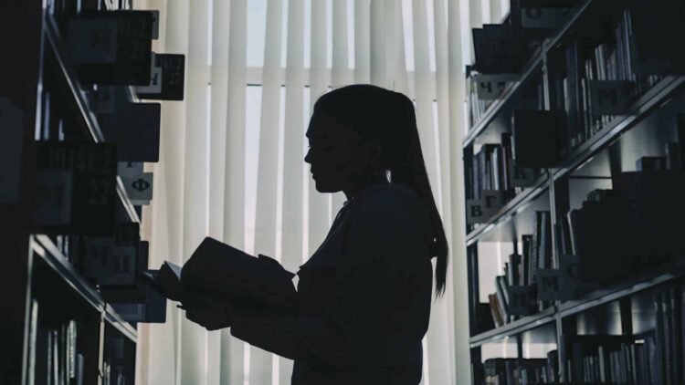 Silhouette of young student girl standing among bookshelves in big library against window holding book turning over pages and reading law student
